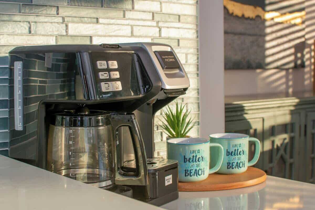 Een koffiezetapparaat op een aanrecht met twee mokken ernaast, een weegschaal en een bakstenen muur op de achtergrond.