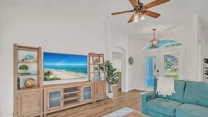 A living room with a large TV, wooden entertainment unit, teal sofa, and a ceiling fan.