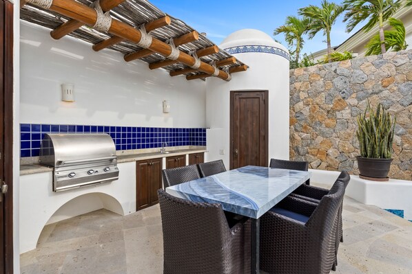 An outdoor kitchen with a grill, dining table, and chairs, surrounded by stone walls and a palm tree. - Casa Estrella 5BR | Infinity Pool, Spa and Ocean Views in Pedregal (Cabo San Lucas)