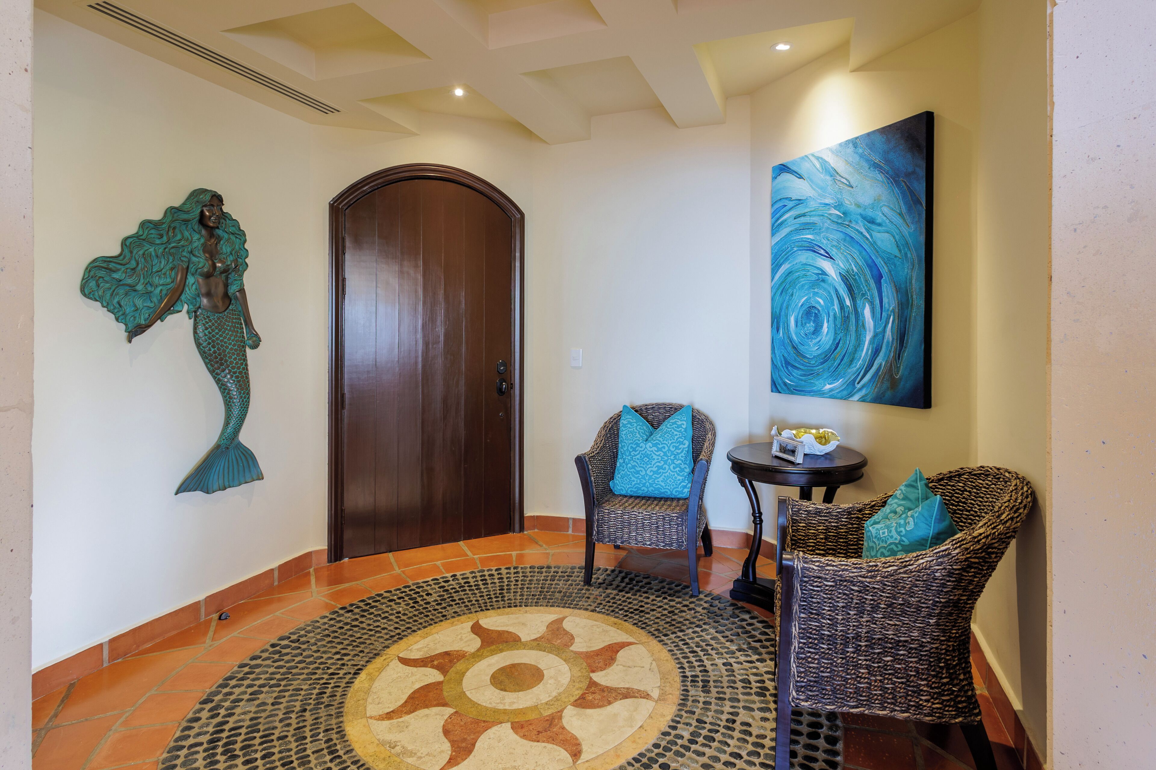 Un pasillo con una puerta de madera, una obra de arte colgada en la pared y dos sillones de mimbre con cojines azules.