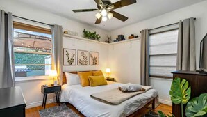 Une chambre à coucher comprenant un lit, des tables de chevet, un ventilateur de plafond, une fenêtre avec des rideaux et une vue sur la verdure à l’extérieur.