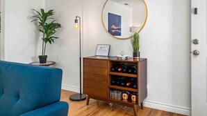 Une pièce avec un canapé bleu, un meuble en bois contenant des bouteilles de vin, une lampe sur pied et un miroir rond accroché au mur.