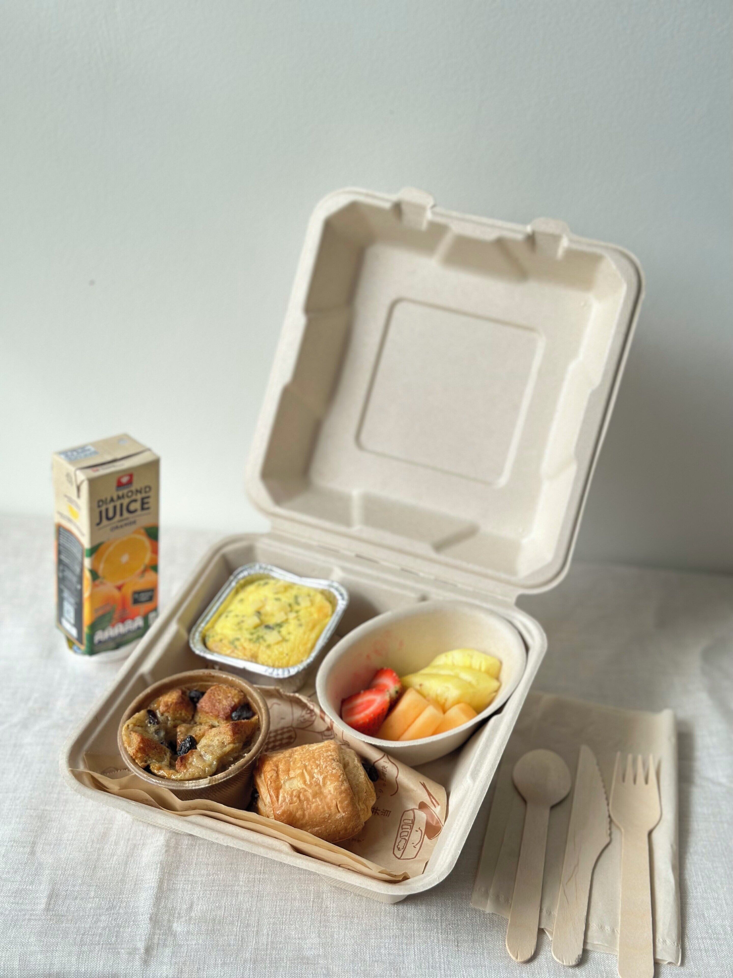 Una comida en un recipiente práctico, con un jugo envasado, un muffin, un bol de fruta y un recipiente pequeño con huevos revueltos.