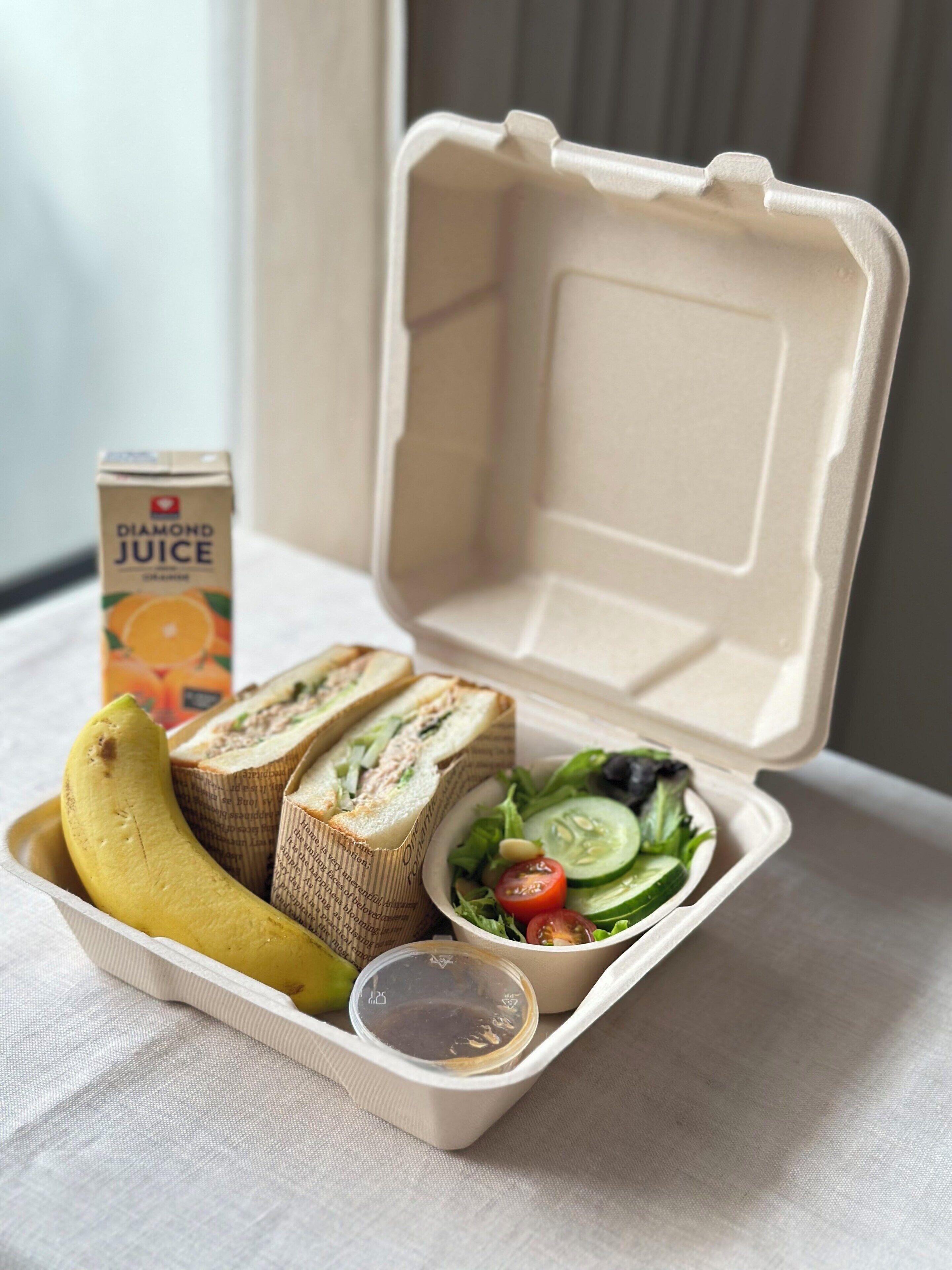 A meal in a takeout container with sandwiches, a banana, a juice carton, and a salad.