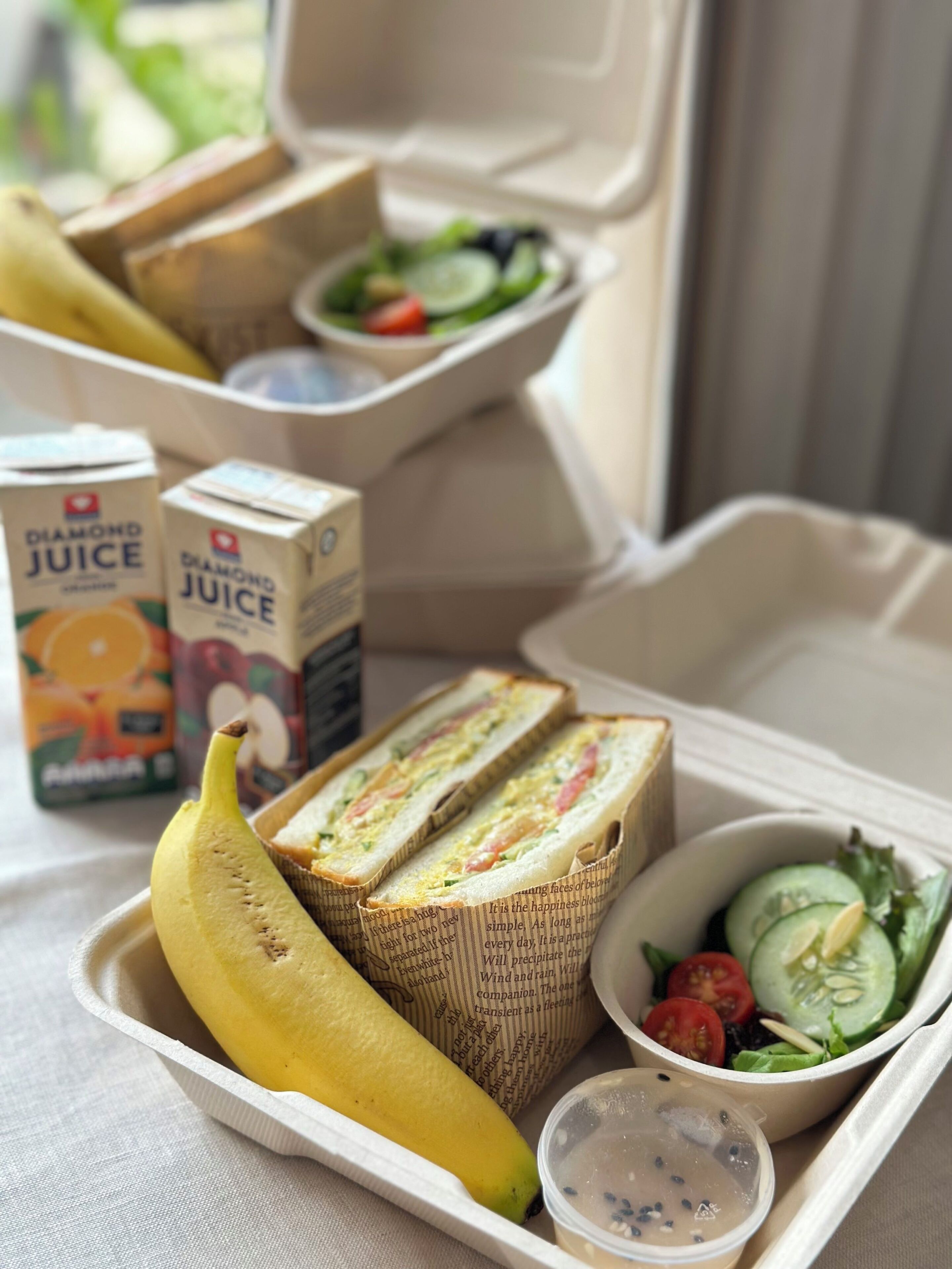 A meal in a takeout container with a sandwich, banana, salad, and juice boxes.