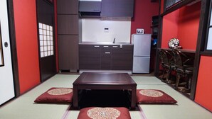 A traditional Japanese-style room with tatami flooring, a low wooden table, and a kitchen area with a refrigerator and cabinets.
