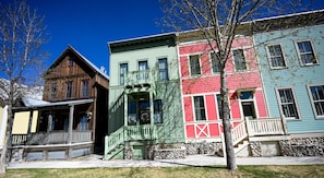 Eine Reihe farbenfroher, historischer Gebäude mit unterschiedlichen Baustilen, darunter ein Holzhaus mit Veranda und ein Gebäude mit grüner Fassade und einem fensterartigen Aufbau wie ein Bauernhaus.