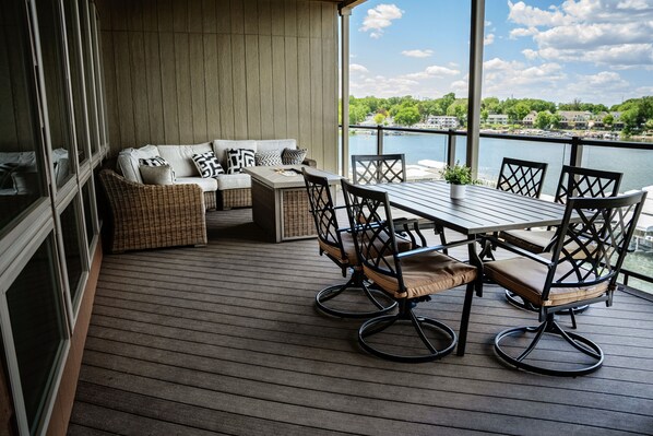 A wooden deck with a dining table and chairs, a sofa with cushions, and a view of a body of water and greenery in the background.