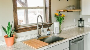 A kitchen with a wooden ceiling, a window with a wooden frame, a sink, a cutting board, a potted plant, and a vase with flowers. - Relaxing, quiet cabin close to Glacier Park! (Columbia Falls)