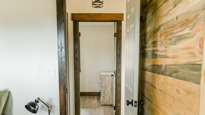 A narrow hallway with wooden flooring, a wooden door on the right, and a bed with a nightstand on the left. - Relaxing, quiet cabin close to Glacier Park! (Columbia Falls)
