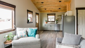 A cozy living room with a grey sofa, a patterned pillow, and a small side table with a plant. A kitchen area with a fridge, sink, and cabinets is visible in the background. - Relaxing, quiet cabin close to Glacier Park! (Columbia Falls)