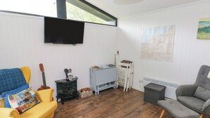 Una sala de estar con un sofá amarillo, un sillón gris, una mesita, una guitarra, un calentador y un televisor montado en la pared.