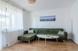 A room with a green sofa, a round white table, a picture on the wall, and a window with sheer curtains.