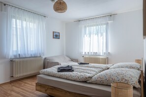 A bedroom with two beds, white curtains, a wooden floor, a radiator, and a small picture on the wall.