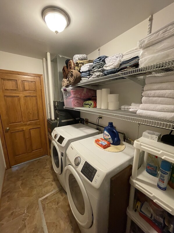 A laundry room with two front-loading washing machines, a dryer, shelves with folded towels, and laundry supplies.