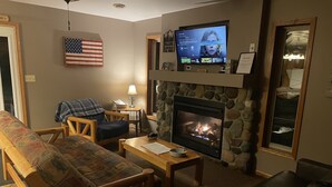 Ein Wohnzimmer mit Kamin aus Stein, einem darüber montierten Fernseher, einem hölzernen Sofa, einem schwarzen Ledersessel und einem gemusterten Sessel.