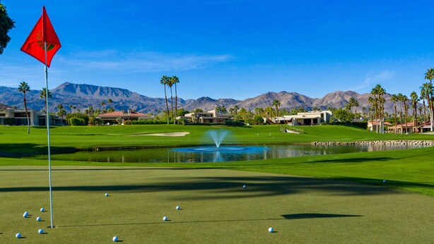A golf course with a flag, a pond, and mountains in the background.
