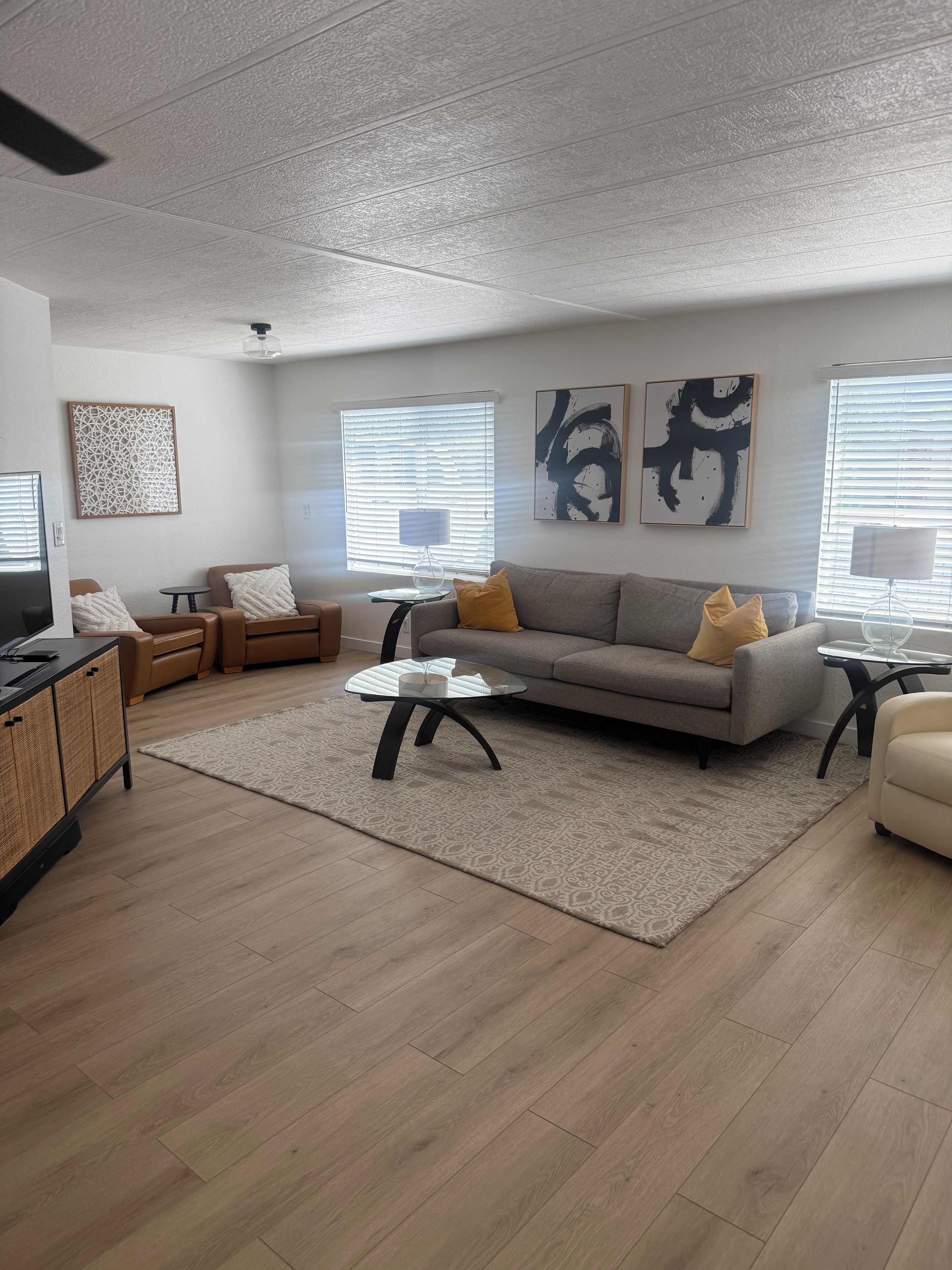 A modern living room with a grey sofa, two armchairs, a coffee table, and a TV. There are abstract wall art pieces and a patterned rug.