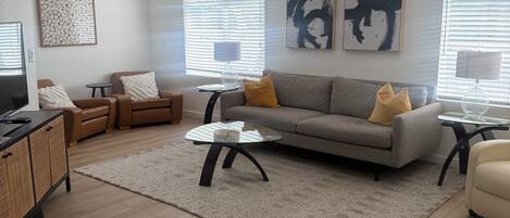 A modern living room with a grey sofa, two armchairs, a coffee table, and a TV. There are abstract wall art pieces and a patterned rug.