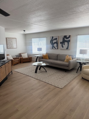 Ein modernes Wohnzimmer mit grauem Sofa, zwei Sesseln, einem Couchtisch und einem Fernseher. An den Wänden befinden sich abstrakte Kunstwerke und ein Teppich mit Muster.