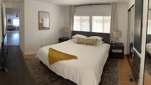 A bedroom with a large bed, a nightstand, a mirror, and a window with blinds.