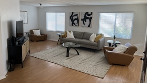 A modern living room with a grey sofa, a coffee table, and a TV on a stand. There are two windows with blinds, a ceiling fan, and a patterned rug on the floor.