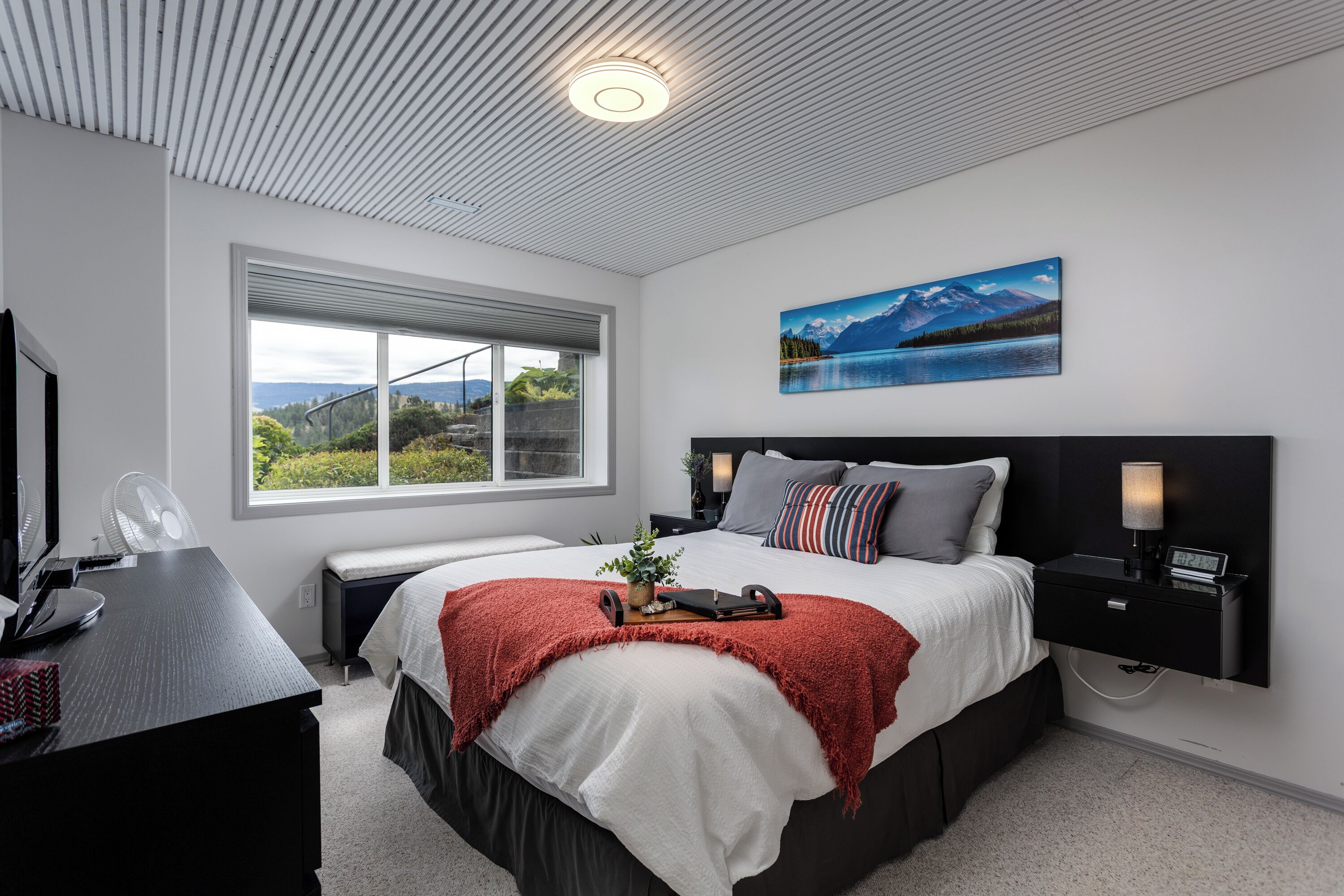 Ein modernes Schlafzimmer mit einem großen Bett, einem Schreibtisch mit Computer, einem Fenster mit Ausblick und einem Bild an der Wand.