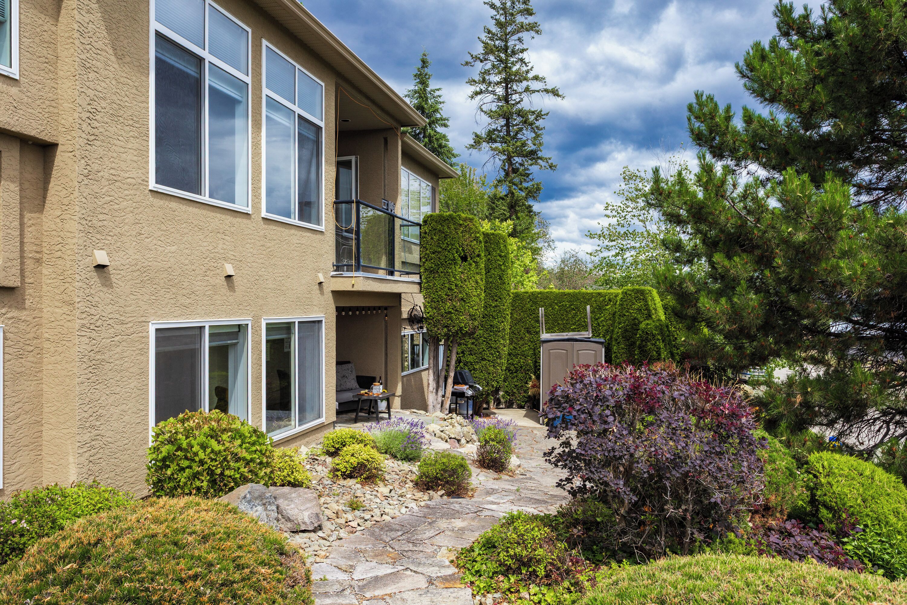 A two-story building with a stone pathway, a balcony, and various shrubs.