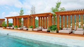 Un área junto a la piscina con pérgolas de madera, asientos acolchados y plantas en macetas.