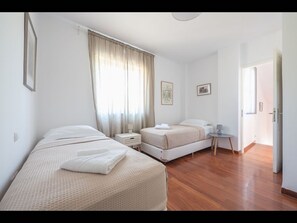 Two single beds with white bedding, bedside tables, and a large window with sheer curtains.