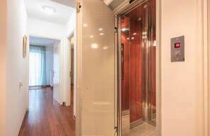 A hallway with a wooden floor, a door leading to an elevator, and a light switch on the wall. - Adelos III Bloom Villa – 5BR 360m² - Sauna - Rooftop (Elliniko)