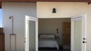 A room with a bed, a small bedside table, and a door leading to a bathroom with a shower. - Huge Views in North Joshua Tree w/ Outdoor Bathtub & Shower (Joshua Tree)