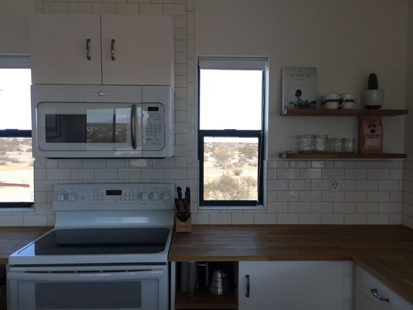 A kitchen with a microwave, oven, and a window with a view. There are shelves with cups and a coffee bag.