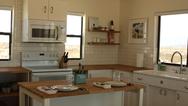 A kitchen with white cabinets, a microwave, a stove, and a wooden countertop. There are shelves with kitchen items and a window with a view. - Huge Views in North Joshua Tree w/ Outdoor Bathtub & Shower (Joshua Tree)