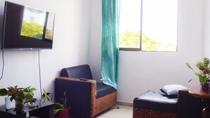 Una habitación pequeña y bien iluminada con un televisor montado en la pared, una sola silla, un sofá y una ventana con cortina.