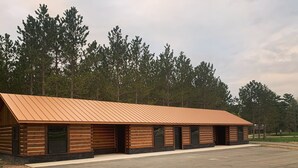 A row of log cabin-style cabins with a metal roof, set in a wooded area with a paved parking lot. - {Big Timbers} Lodging next to North Entrance of Itasca State Park (Shevlin)