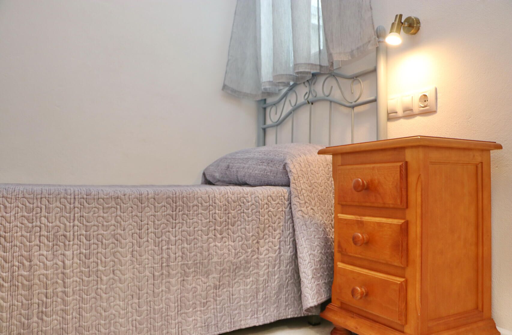 Ein Schlafzimmer mit einem Bett, einer Holzkommode, einer Steckdose und einem Fenster mit durchsichtigem Vorhang.