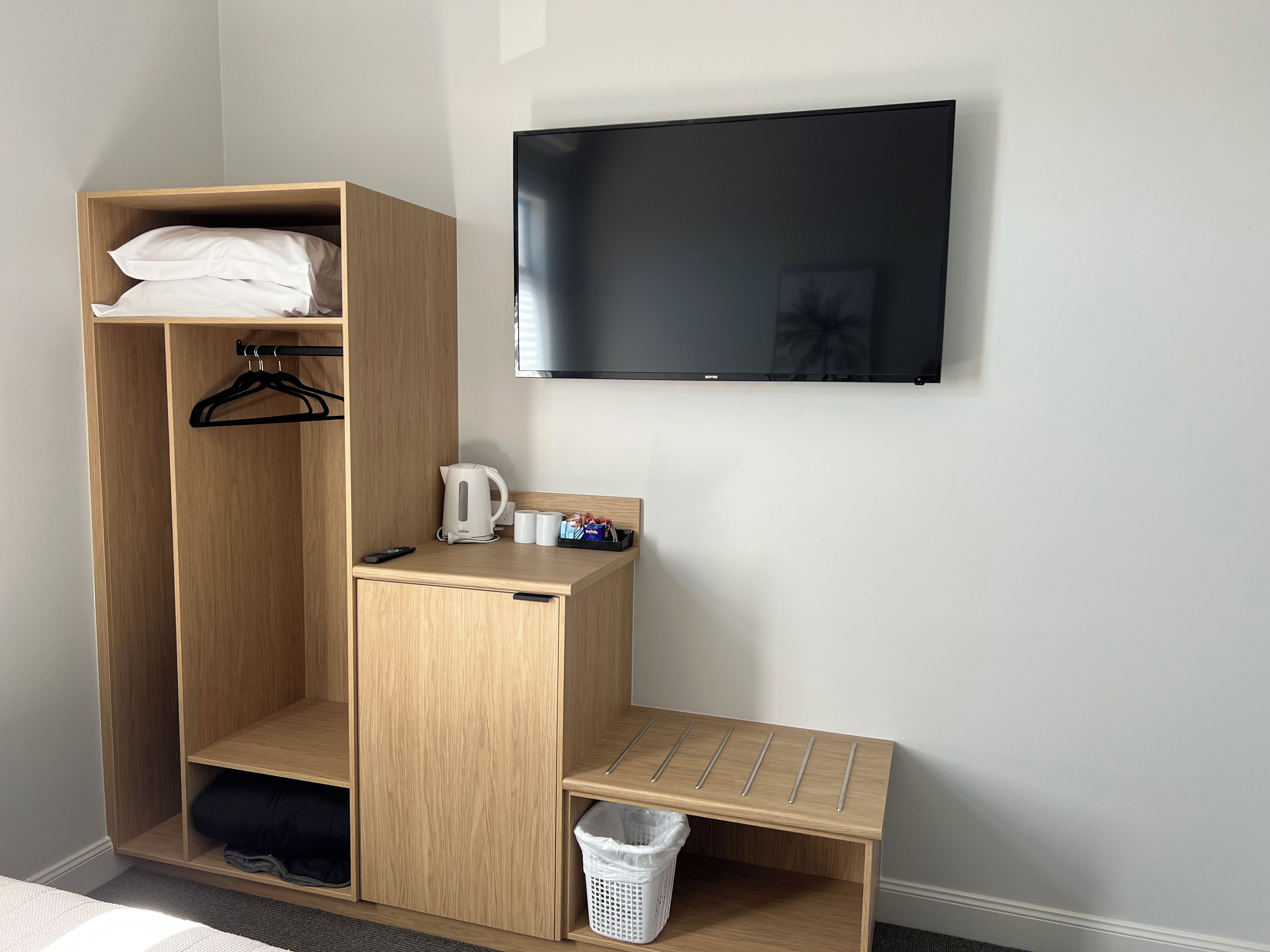 Een hotelkamer met een houten garderobe, een flatscreen-tv aan de muur, een klein tafeltje met waterkoker en bekers, en een prullenbak.