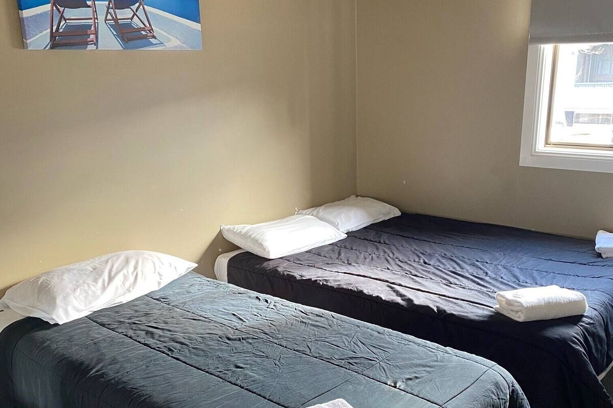 Two single beds with dark bedding and white pillows, a window with blinds, and a painting of a beach scene on the wall.