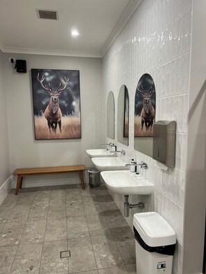 A bathroom with three sinks, a bench, and a trash can.