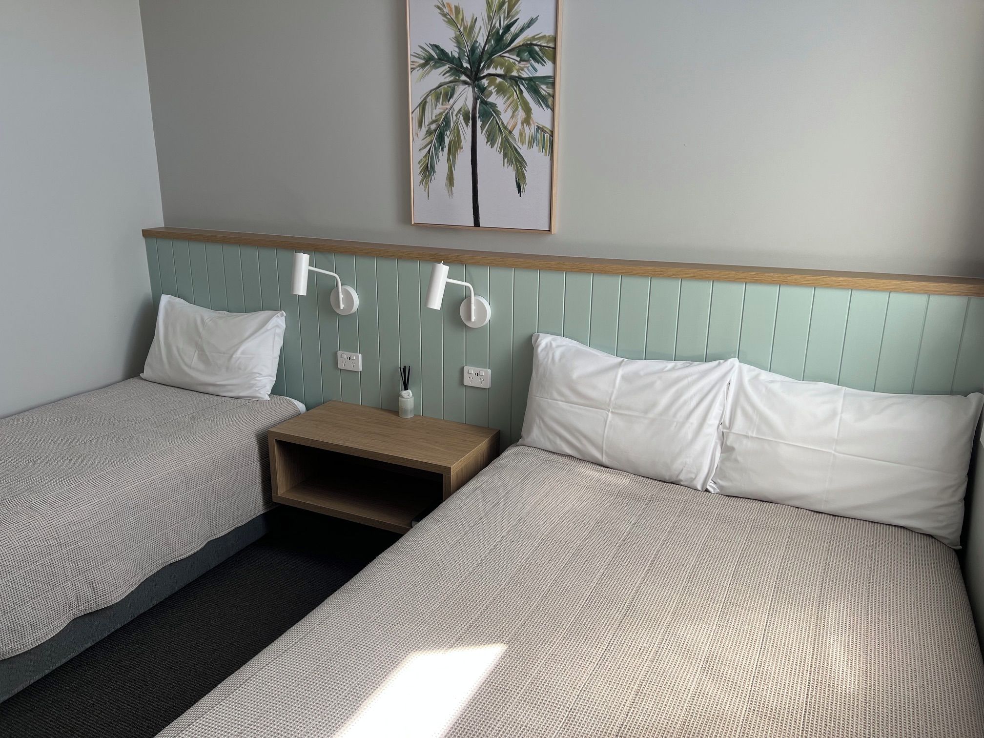 Een hotelkamer met twee bedden, een nachtkastje, een wandlamp en een schilderij van een palmboom.