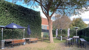 Un patio al aire libre con bancos y mesas de madera, rodeado por un seto y árboles.