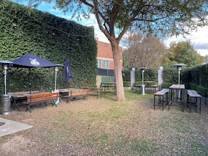 Un patio al aire libre con bancos y mesas de madera, rodeado por un seto y árboles.