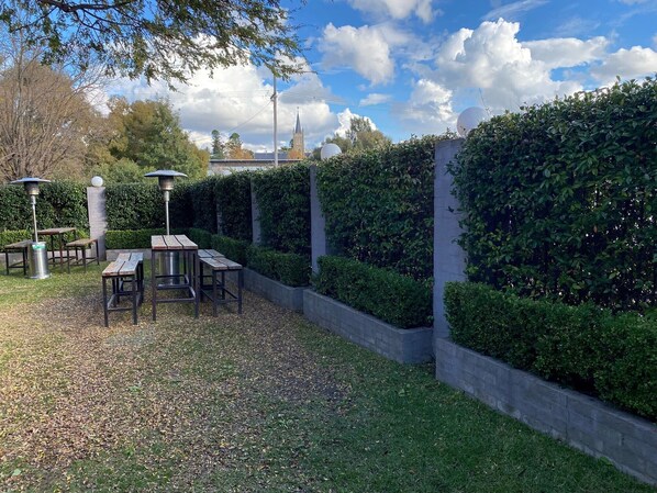 Un patio al aire libre con mesas y sillas, rodeado de setos cuidados y un muro de piedra.