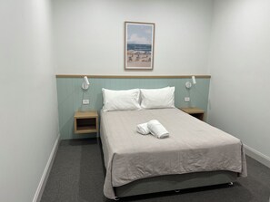 A hotel room with a bed, two bedside tables, a wall-mounted lamp, a power outlet, and a framed picture of a beach scene.