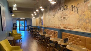 Interior de un restaurante con mesas de madera y sillas amarillas, una pared con la señalización 'Shamrock Inn' y vista de la calle.