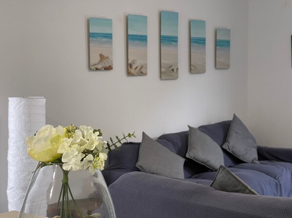 A living room with a grey sofa, a vase with white flowers, and a series of beach-themed paintings on the wall.