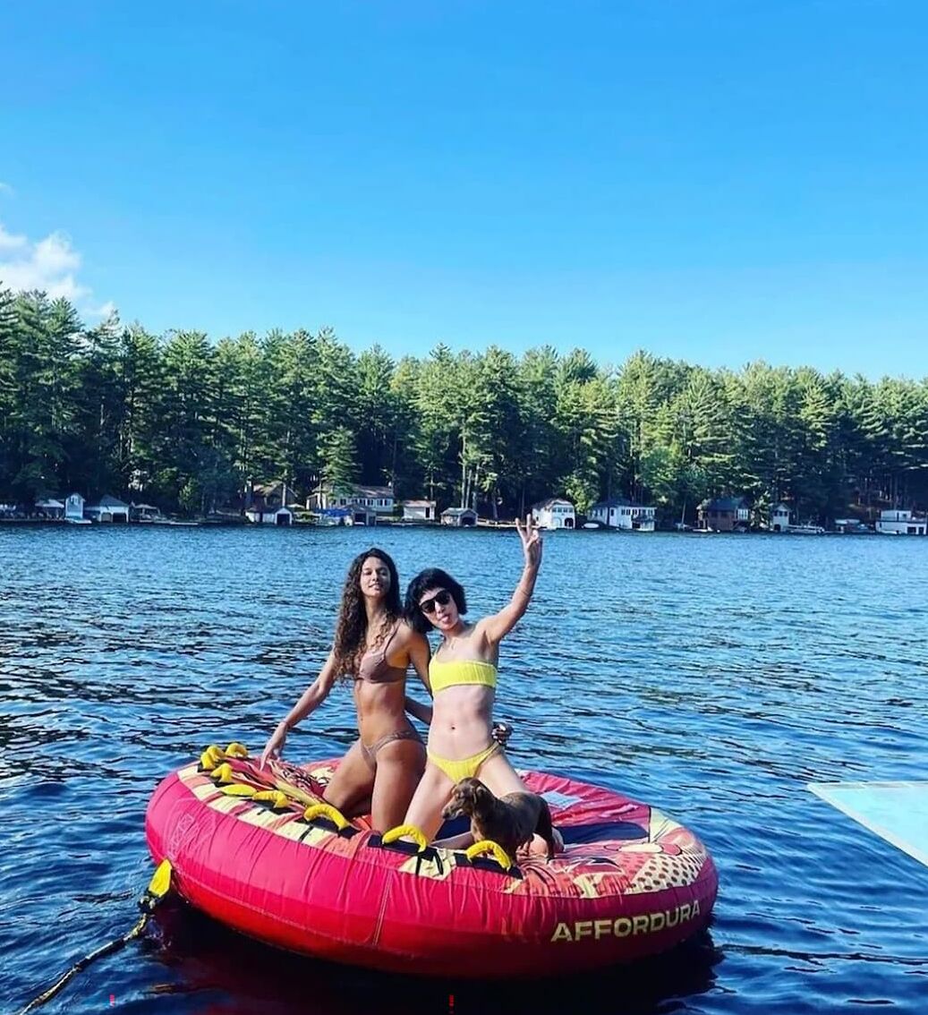 Two people on a red inflatable raft with a dog, surrounded by water and a forested shoreline.