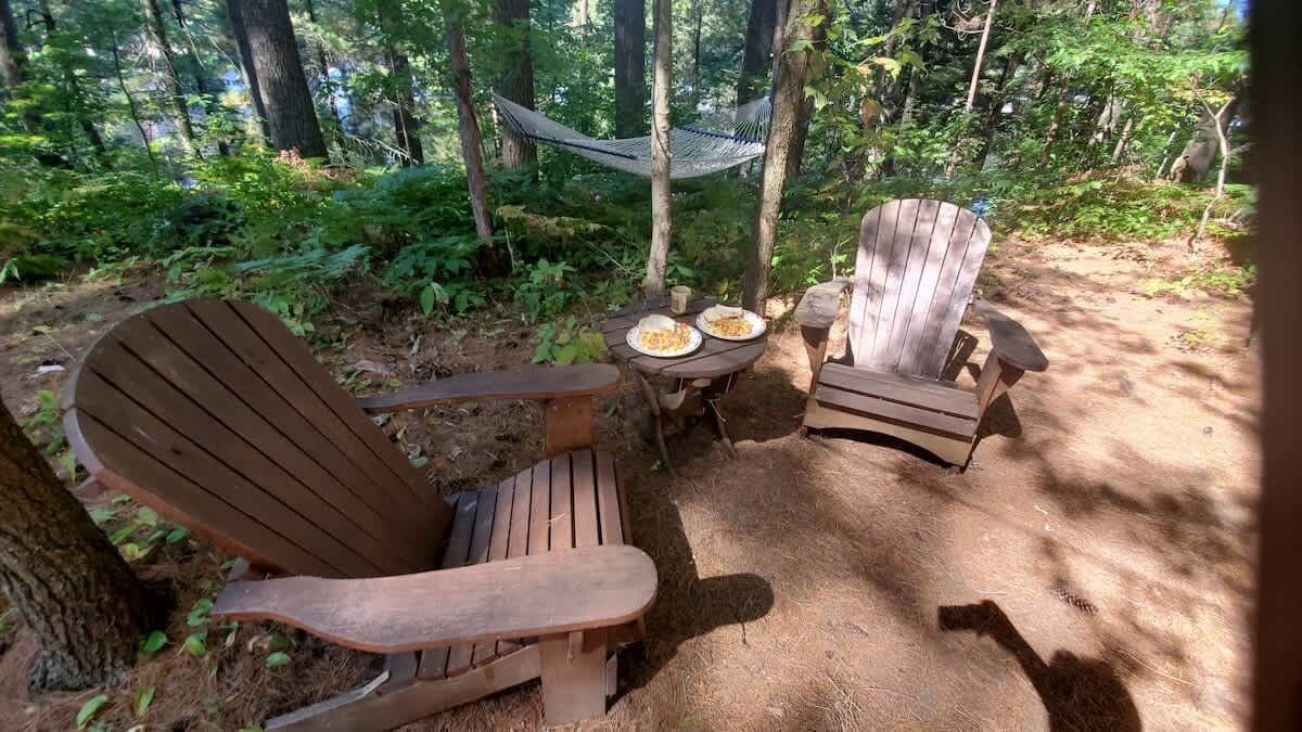 A forested area with two wooden Adirondack chairs and a small table with plates and cups.