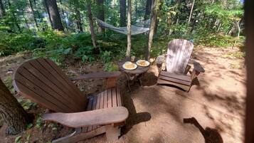 A forested area with two wooden Adirondack chairs and a small table with plates and cups.